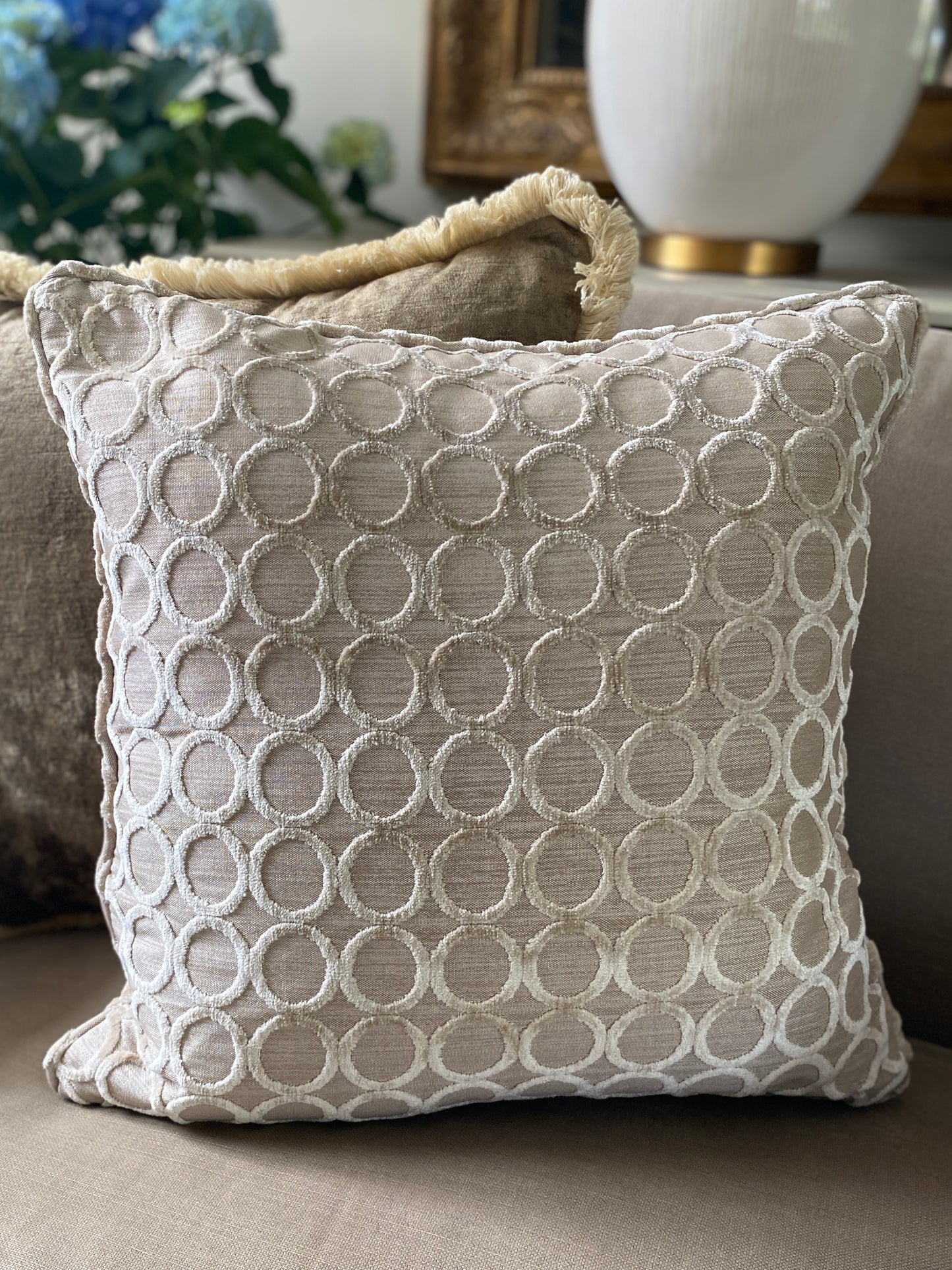 Taupe square cushion with beige background and cut pile circle velvet design with piping in front of brown velvet cushion with fringing.