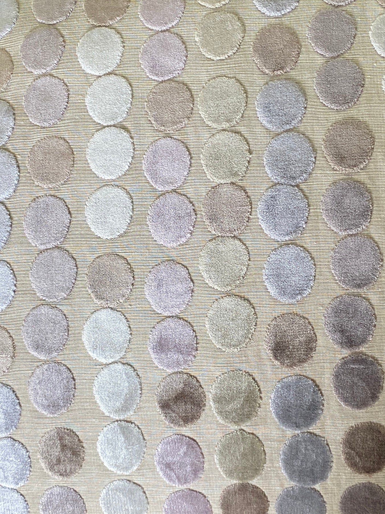 Close up of pastel dots in lilac pink, silvery grey, mink, pale gold and off white on cut pile velvet cushion with a beige background.
