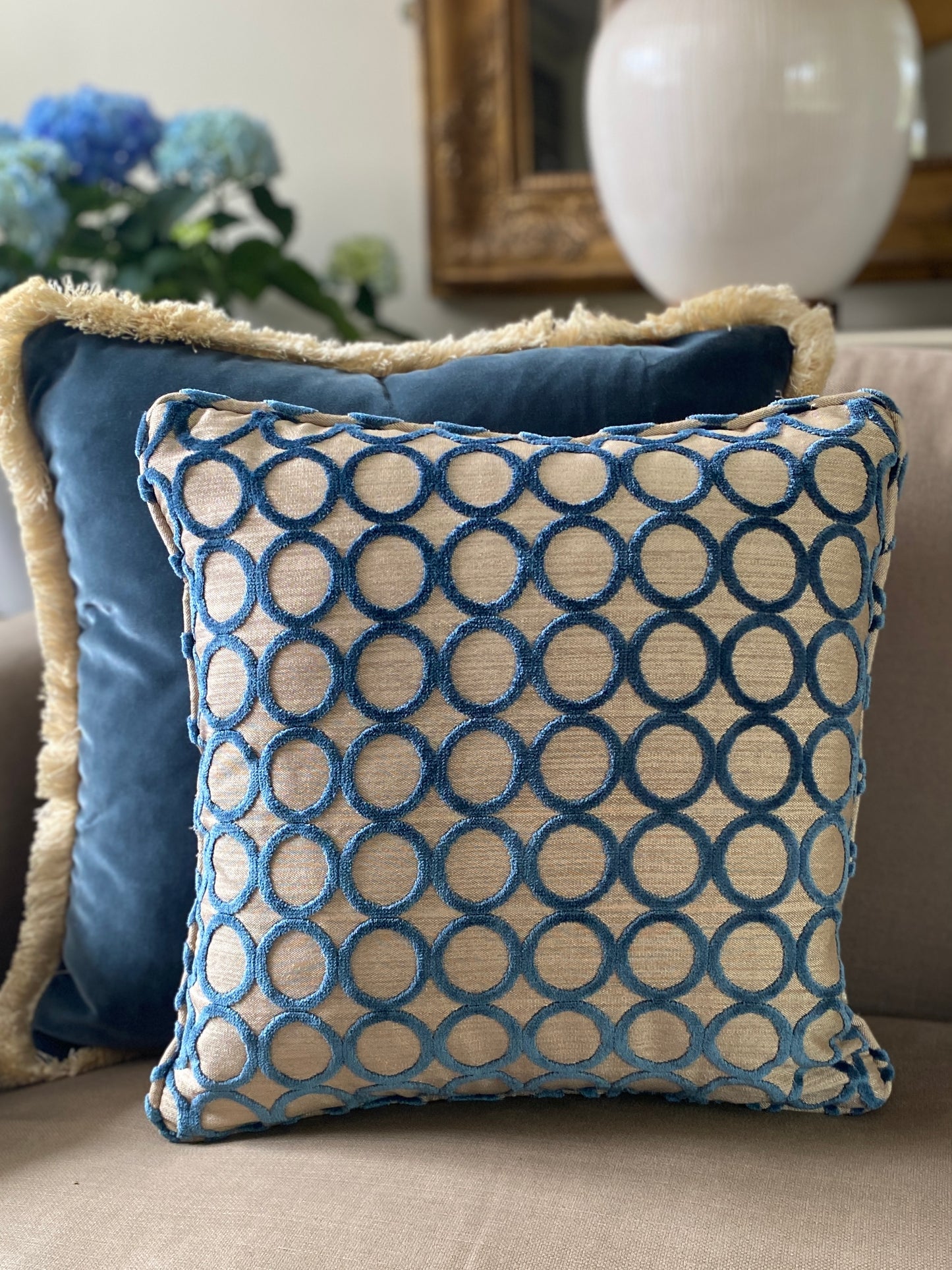Square cut pile velvet cushion with self piping in teal with circle design and beige background. behind it teams a teal velvet larger cushion with fringing.