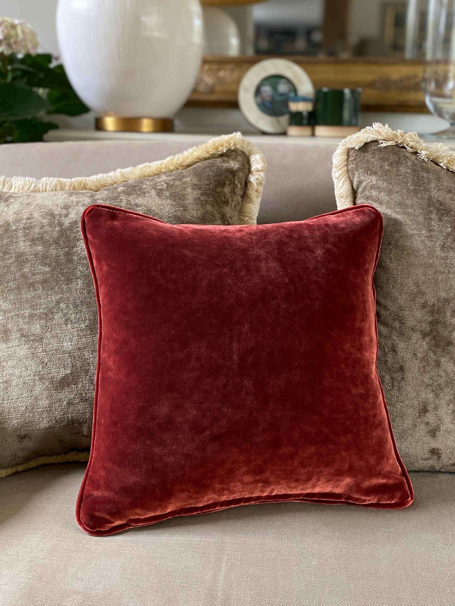 square velvet cushion in autumnal chestnut brown with piping around the edge. Is sat with two brown vintage feel larger cushions with fringing.