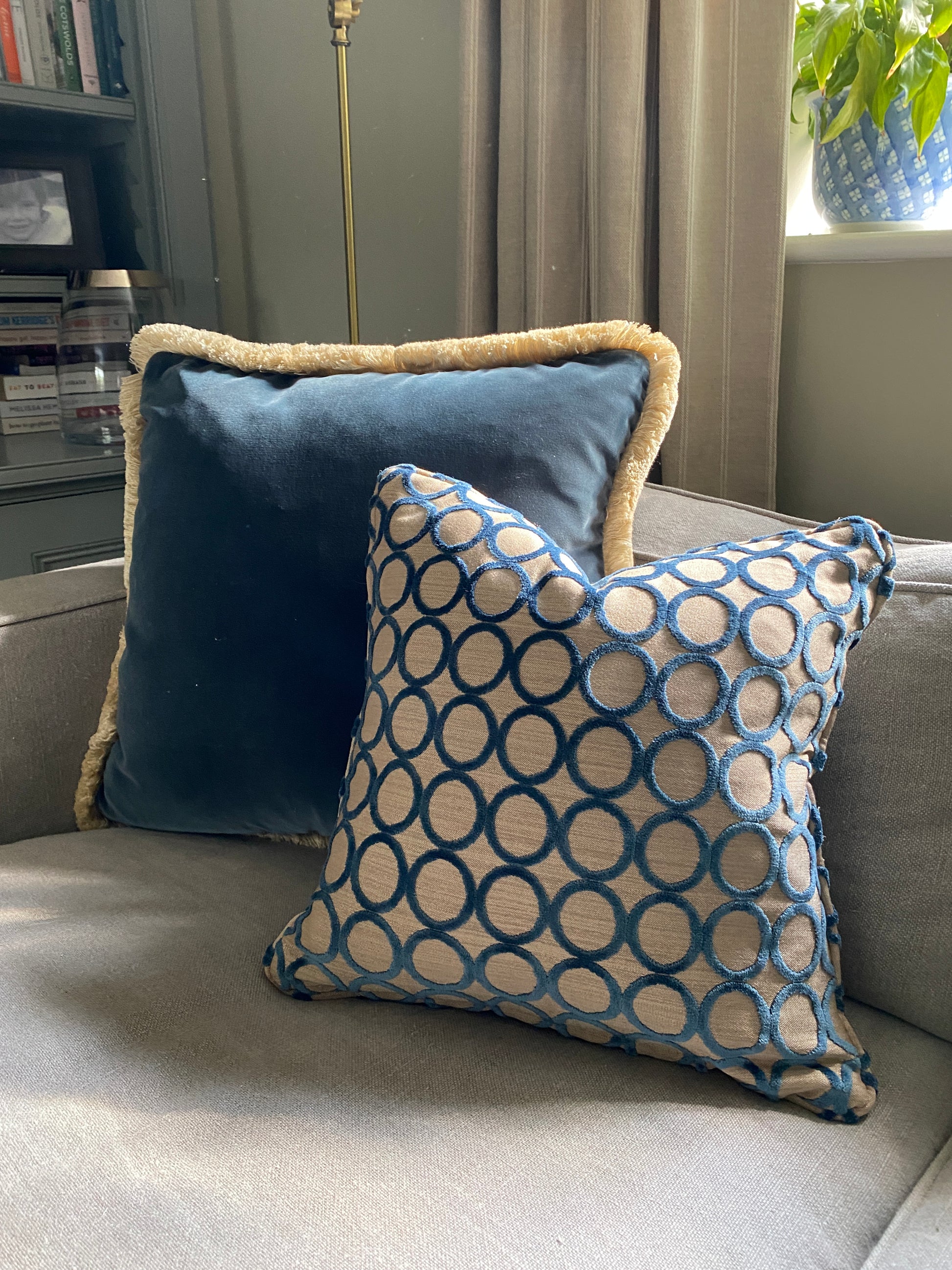 Square cut pile velvet cushion with self piping in teal with circle design and beige background. behind it teams a teal velvet larger cushion with fringing.