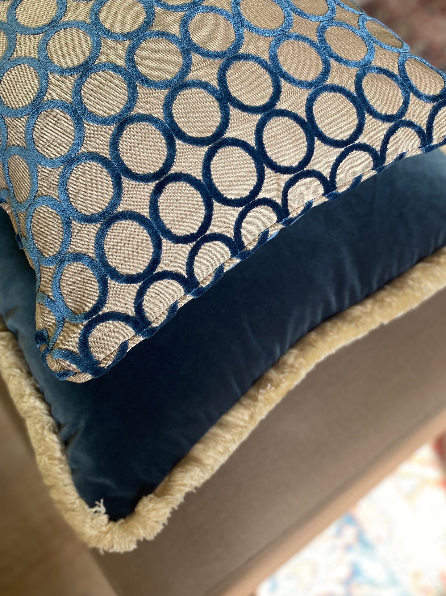 Square cut pile velvet cushion with self piping in teal with circle design and beige background.  Underneath it teams a teal velvet larger cushion with fringing.