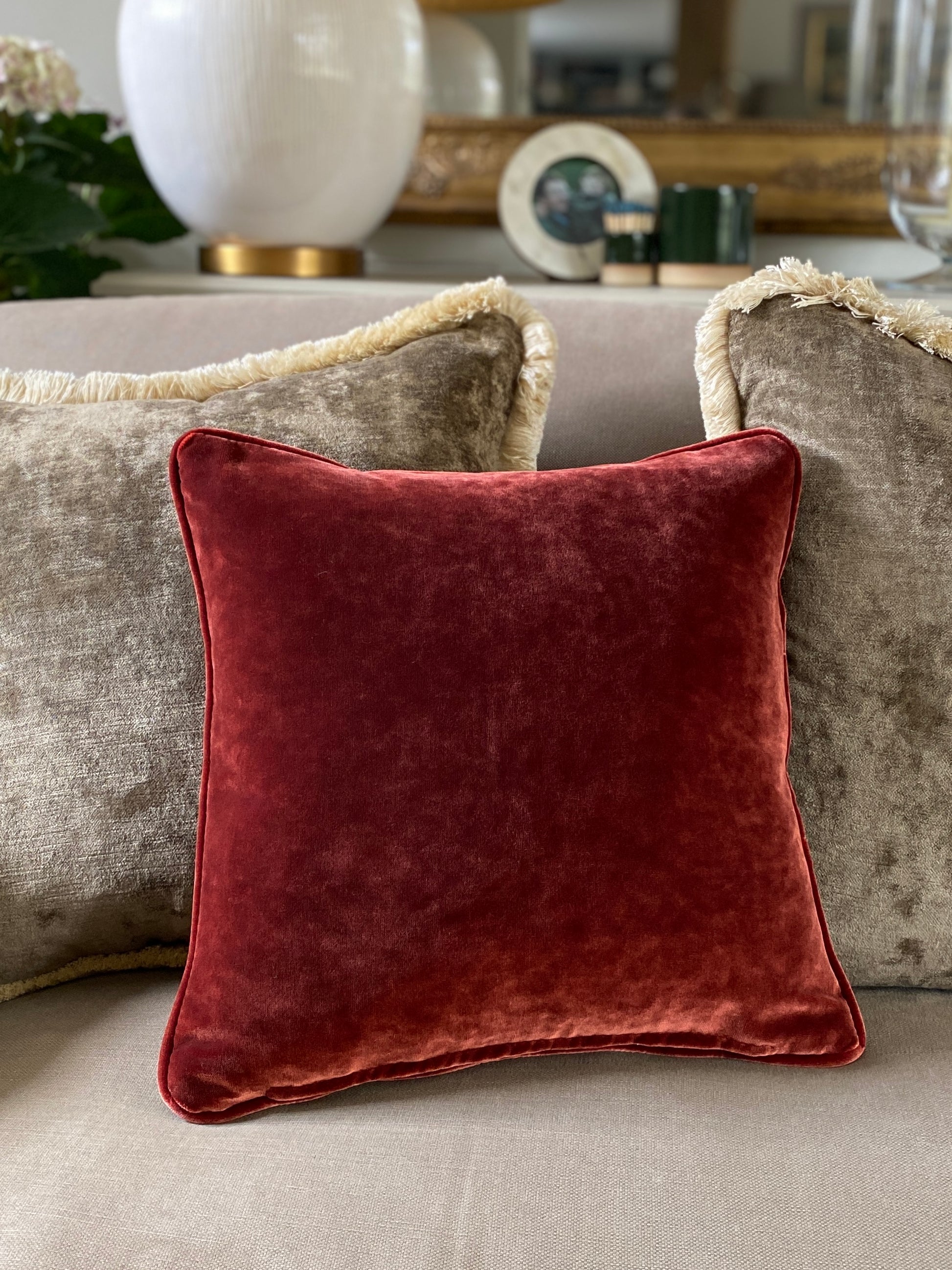 square velvet cushion in autumnal chestnut brown with piping around the edge. Is sat with two brown vintage feel larger cushions with fringing.