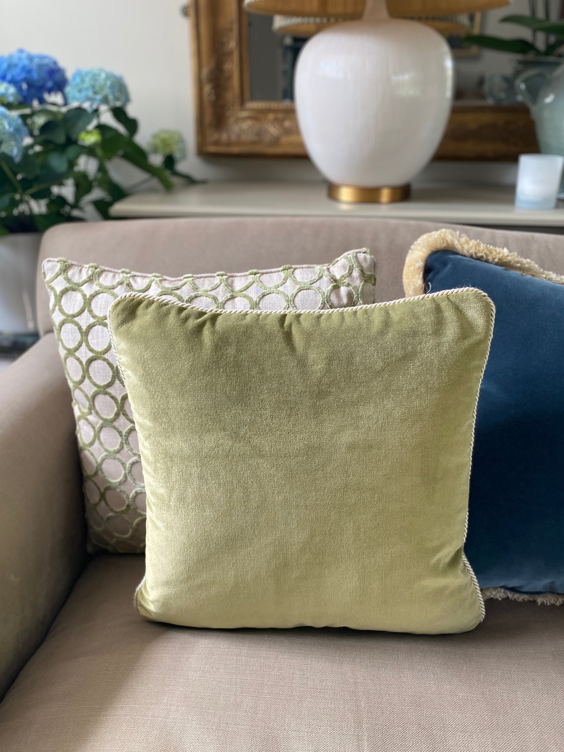 Moss green velvet square cushion with natural cord trim in front of a velvet circle design green cushion and a teal velvet cushion with fringing.