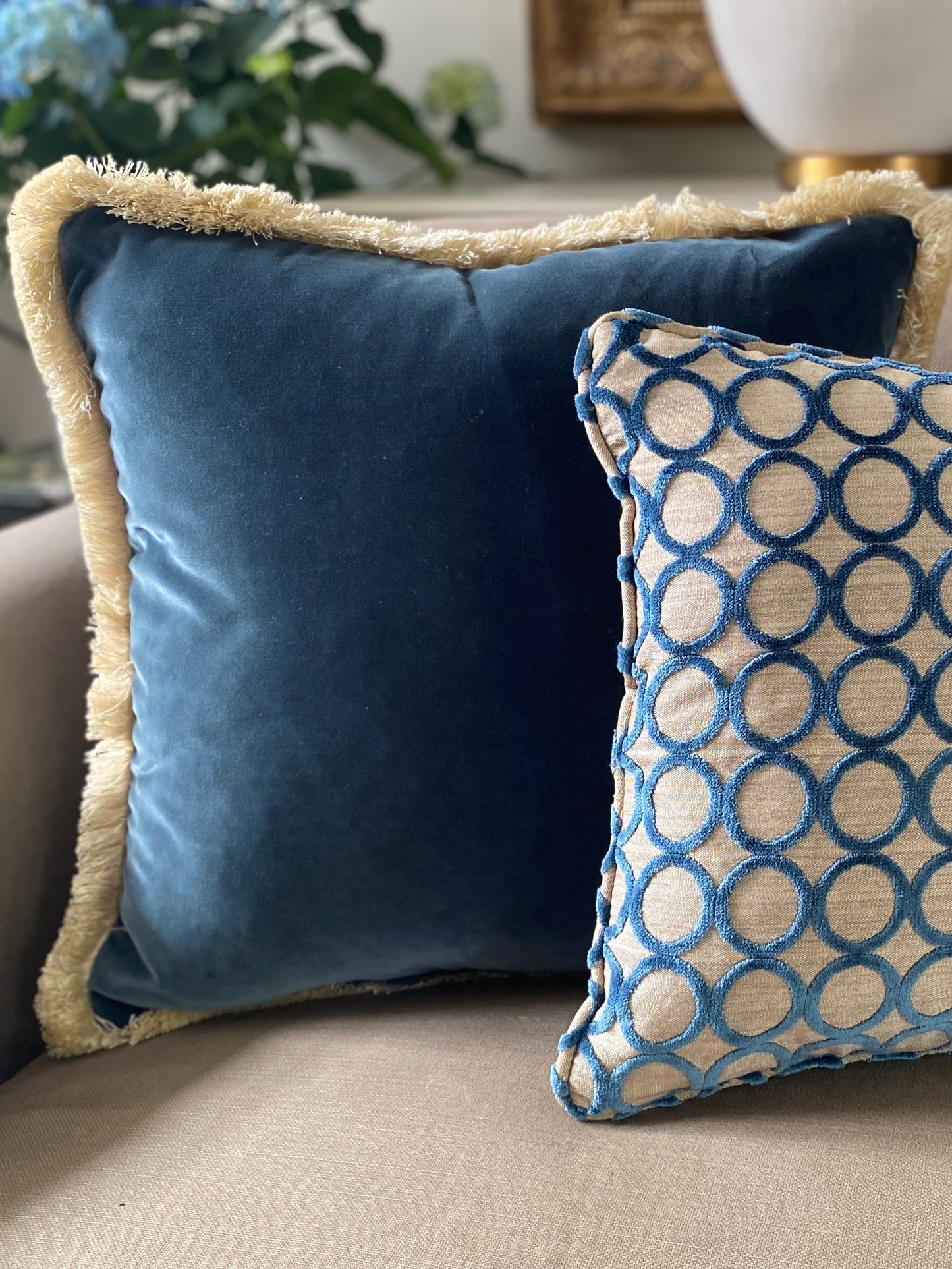 Square cut pile velvet cushion with self piping in teal with circle design and beige background. Behind it teams a teal velvet larger cushion with fringing.