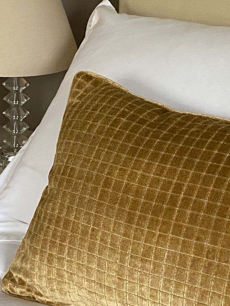 Gold velvet cushion on a bed with white pillows and a lamp in the background